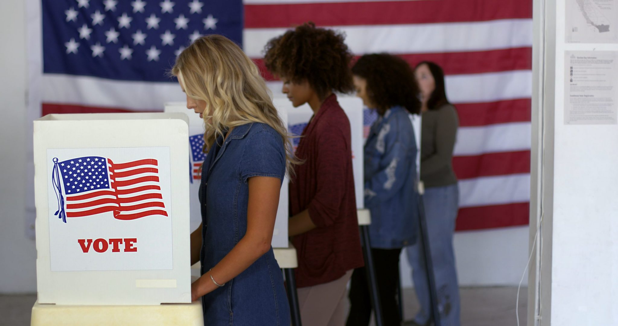 Four women of various demographics filling in ballots and casting votes in booths at polling station, US flag on wall at back.